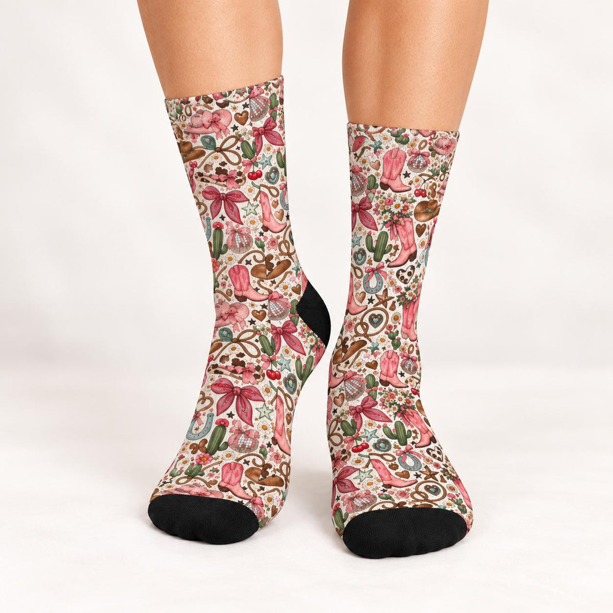 Front view of retro cowgirl socks worn against a clean white studio background. Detailed pink and green western print features cowboy boots, oversized bows, wildflowers, hearts, and horseshoes with black toe accents.