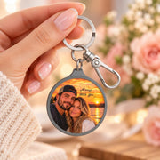 Personalized round photo keychain held in hand, featuring a custom couple picture at sunset with silver key ring and clip hardware.