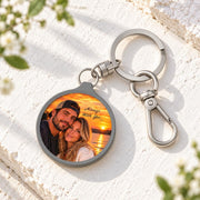 Round photo keychain displayed on a light fabric surface with greenery, featuring a personalized couple image and polished silver hardware.