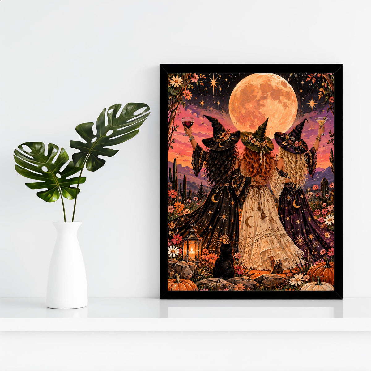 Funky witchy printable displayed in a black frame against a clean white wall beside a small green plant. The artwork features three witches in black, cream, and purple gowns beneath a peach moon, surrounded by wildflowers.