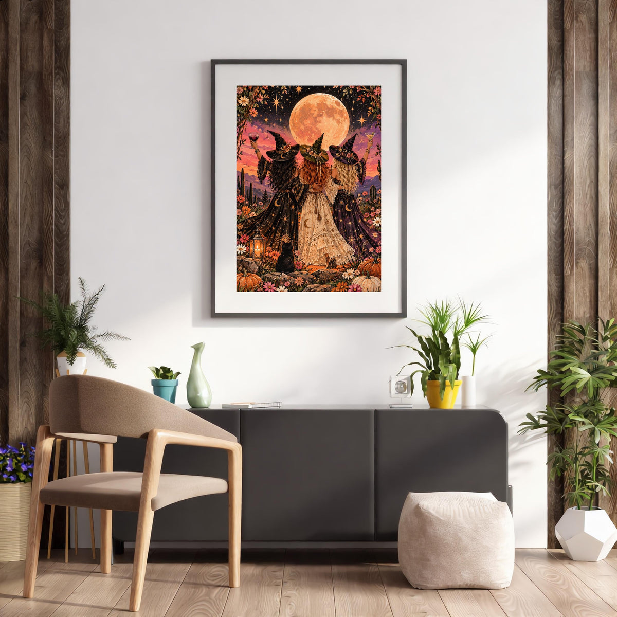 Framed witchy fantasy art displayed above a wooden desk and chair in a bright workspace. The colourful print features three women wearing decorated witch hats, raising cocktails beneath a magical star-filled sky.