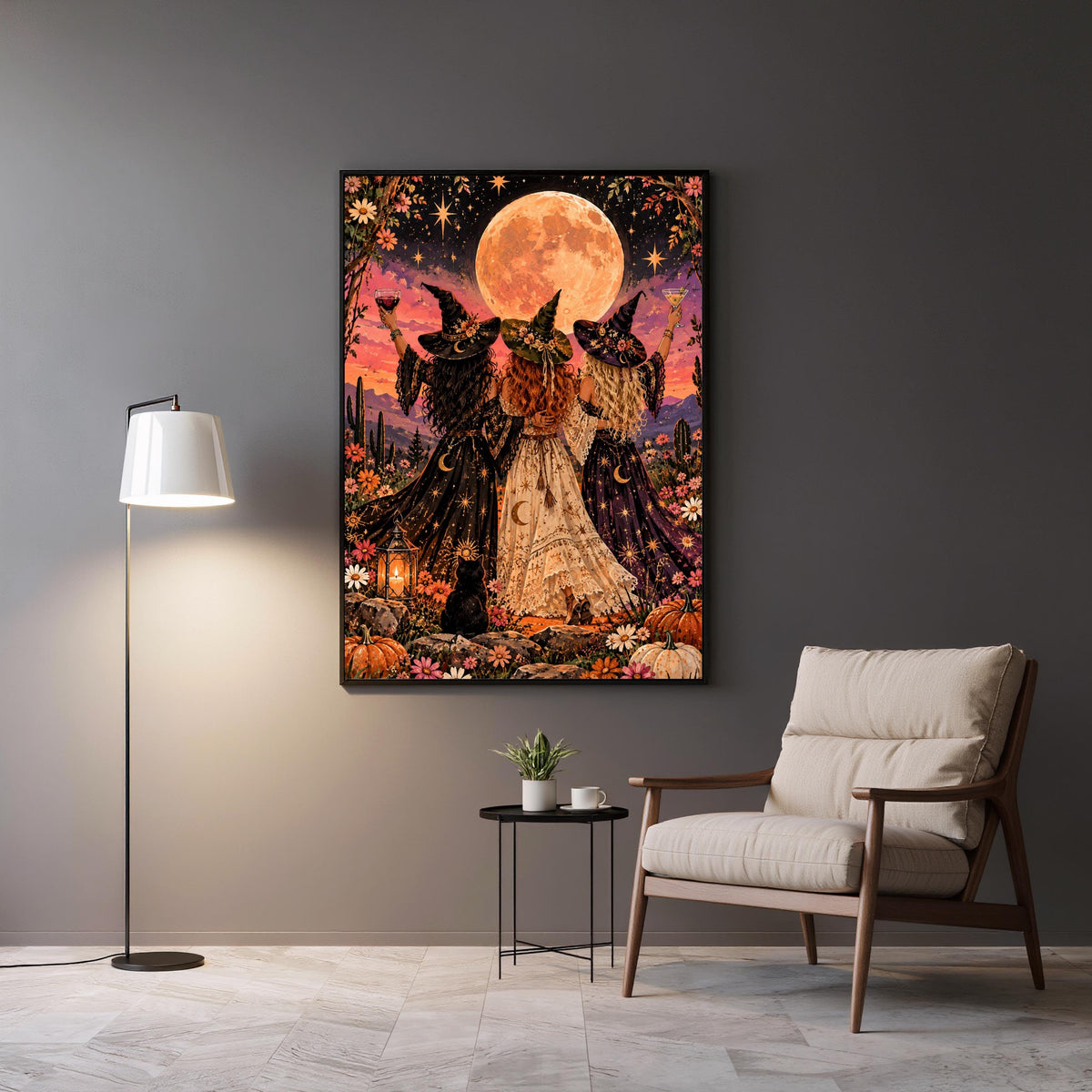 Framed witchy boho wall art displayed above a grey accent chair and side table. The artwork features three magical women celebrating beneath a peach-coloured moon, surrounded by stars and wildflowers.