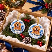 Two personalized ceramic golden retriever ornaments featuring Cooper's name, year 2015, and blue watercolor backgrounds, presented in a holiday gift box with navy ribbon, evergreen branches, and festive berries.