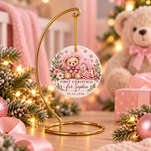 Personalized Baby’s First Christmas ornament in a pink teddy bear and rocking horse design, displayed in a soft holiday scene with ornaments, greenery, and warm lights.