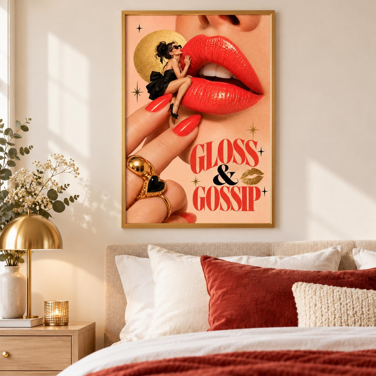 Gloss and Gossip retro lips wall art displayed in a gold frame above a cream bed with rust-coloured pillows. The glamorous poster features oversized coral-red lips, a miniature woman in a black dress, gold jewellery, and vintage typography against a peach background.