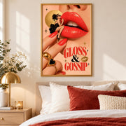 Gloss and Gossip retro lips wall art displayed in a gold frame above a cream bed with rust-coloured pillows. The glamorous poster features oversized coral-red lips, a miniature woman in a black dress, gold jewellery, and vintage typography against a peach background.