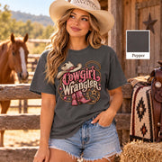 Woman wearing a washed charcoal pepper T-shirt with pink Cowgirl Wrangler artwork, Western boots, cowboy hat and horseshoe graphics, styled with denim shorts at a horse ranch.