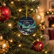 Personalized Army family ornament hanging on a lit Christmas tree beside same-size red and gold baubles, showing a soldier on a snowy ridge under green northern lights and the example lettering Proud Army Mom, Ethan, Christmas 2026