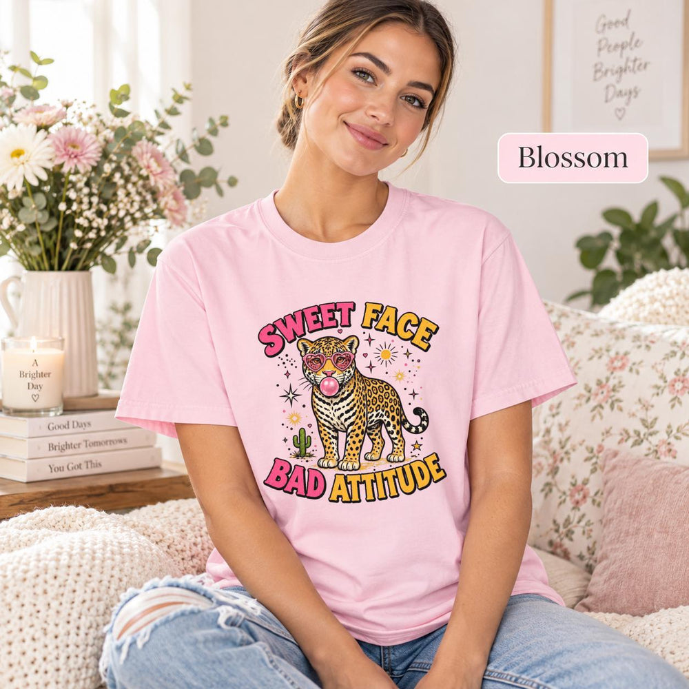 Woman sitting on a sofa wearing a Blossom pink T-shirt with a retro leopard, heart sunglasses, bubblegum, and Sweet Face Bad Attitude lettering.