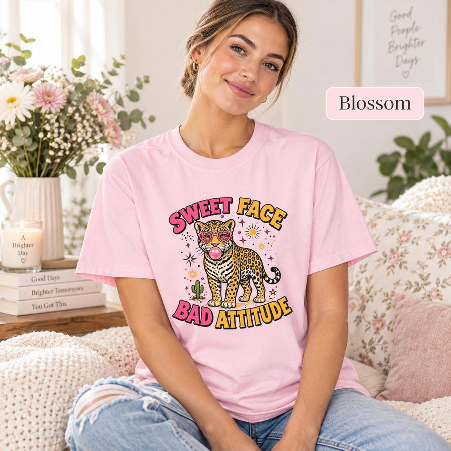 Woman sitting on a sofa wearing a Blossom pink T-shirt with a retro leopard, heart sunglasses, bubblegum, and Sweet Face Bad Attitude lettering.