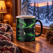 Steaming black colour-changing loon mug on a cabin windowsill beside a knit blanket and pine cones, showing a loon on a dark lake under green northern lights, with snowy spruce trees outside