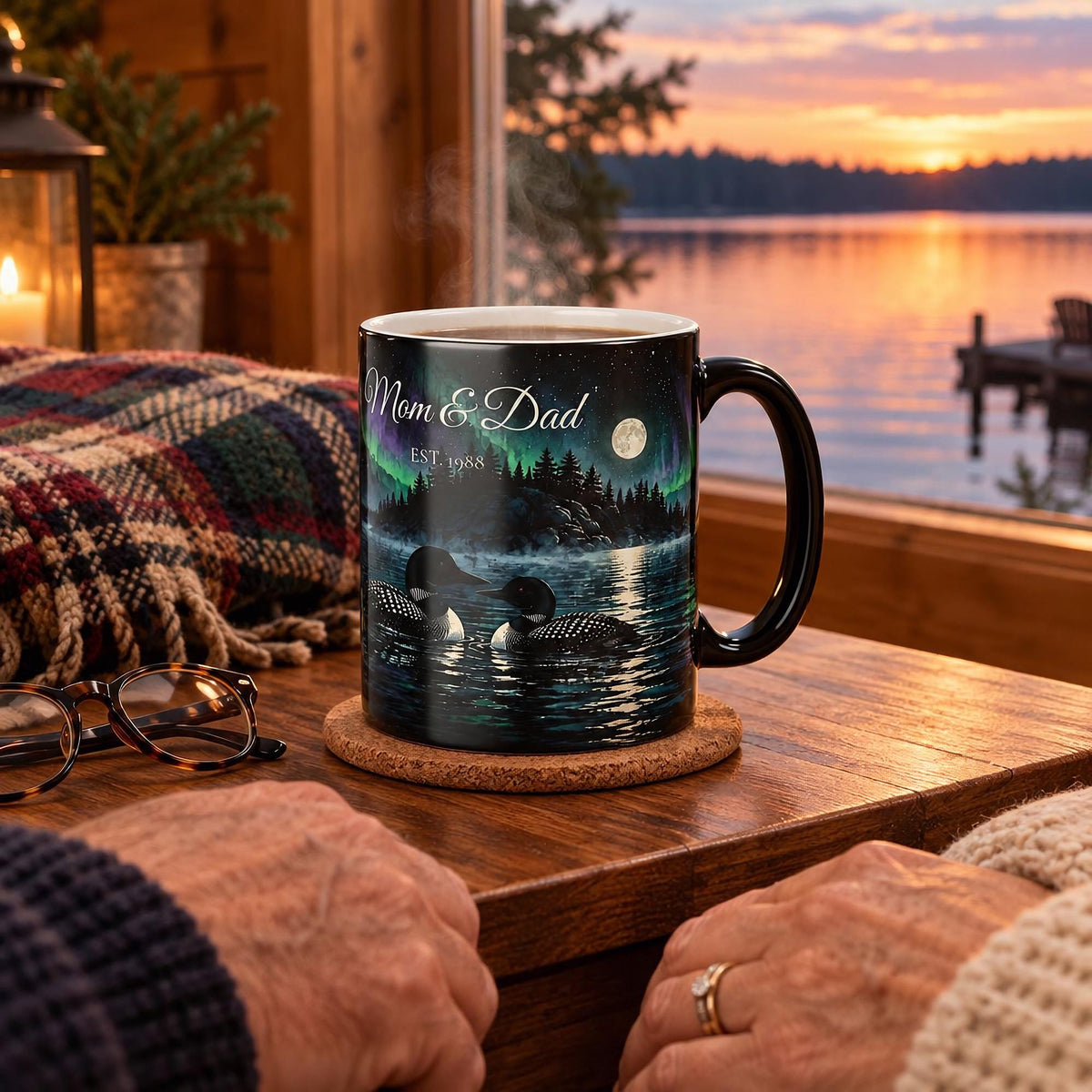 Personalized loon couple mug on a wooden side table by a cottage window overlooking a lake at dusk, with reading glasses, a plaid blanket and an older couple holding hands nearby